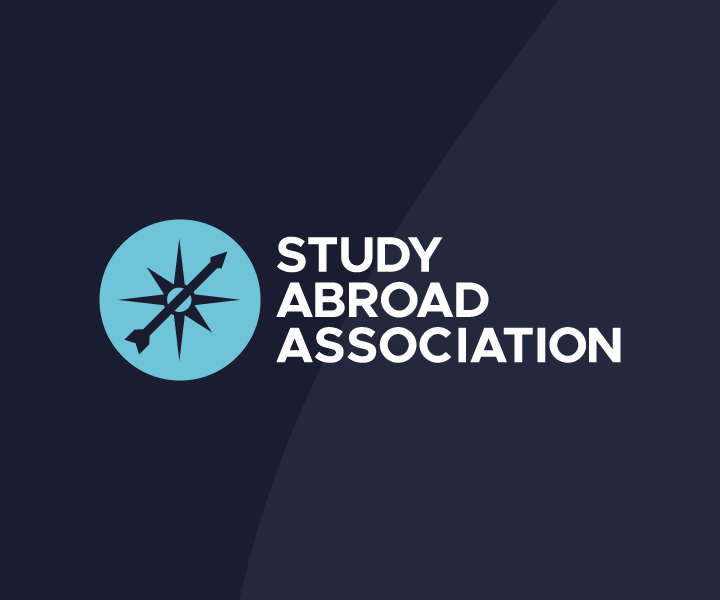 The Study Abroad Association logo set against a dark navy blue background with a subtle diagonal color split. On the left, there is a light blue circular icon containing a dark compass rose with an arrow pointing toward the upper right. To the right of the icon, the words "STUDY ABROAD ASSOCIATION" are written in a bold, white, sans-serif font stacked in three lines.