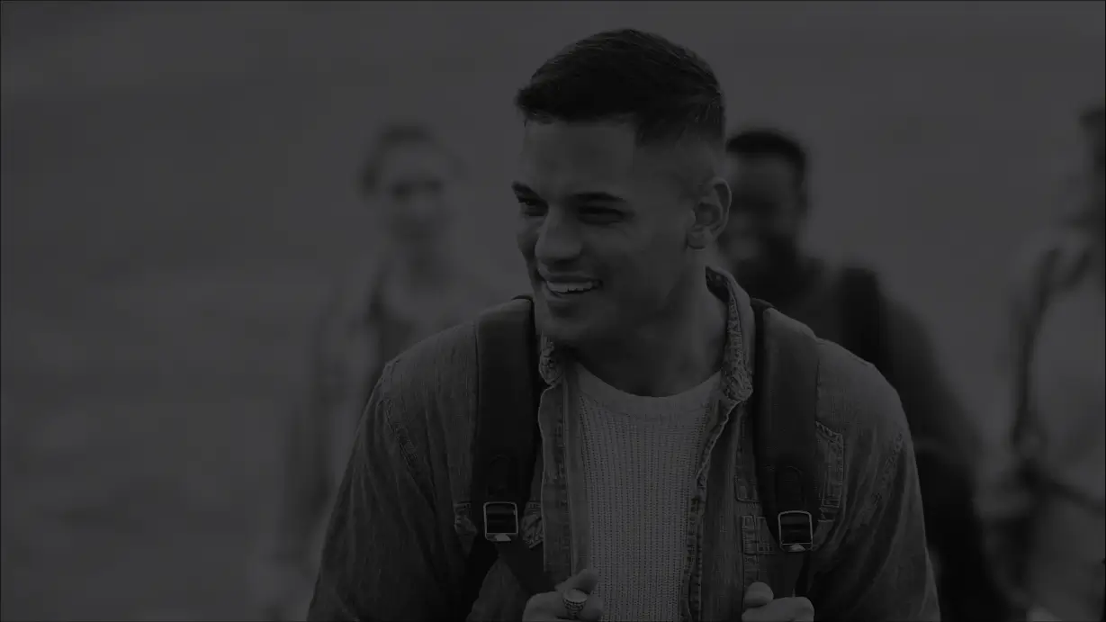 A monochrome, dark-toned photograph of a smiling young man with a short haircut, wearing a backpack and a denim shirt over a sweater, looking slightly to his left. Other blurred figures appear in the background, suggesting a group setting or a hike.