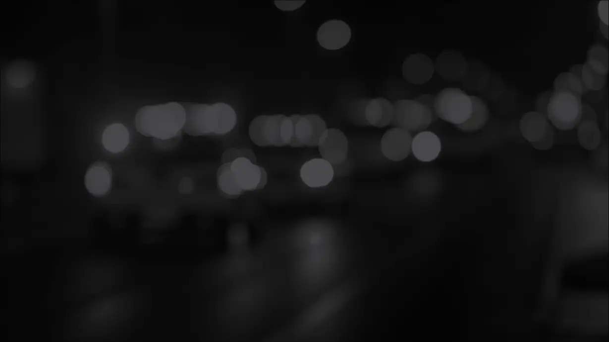 A dark, out-of-focus, black-and-white image of a street at night. Soft, blurry bokeh circles from streetlights and vehicle headlights illuminate a dark roadway in the background.