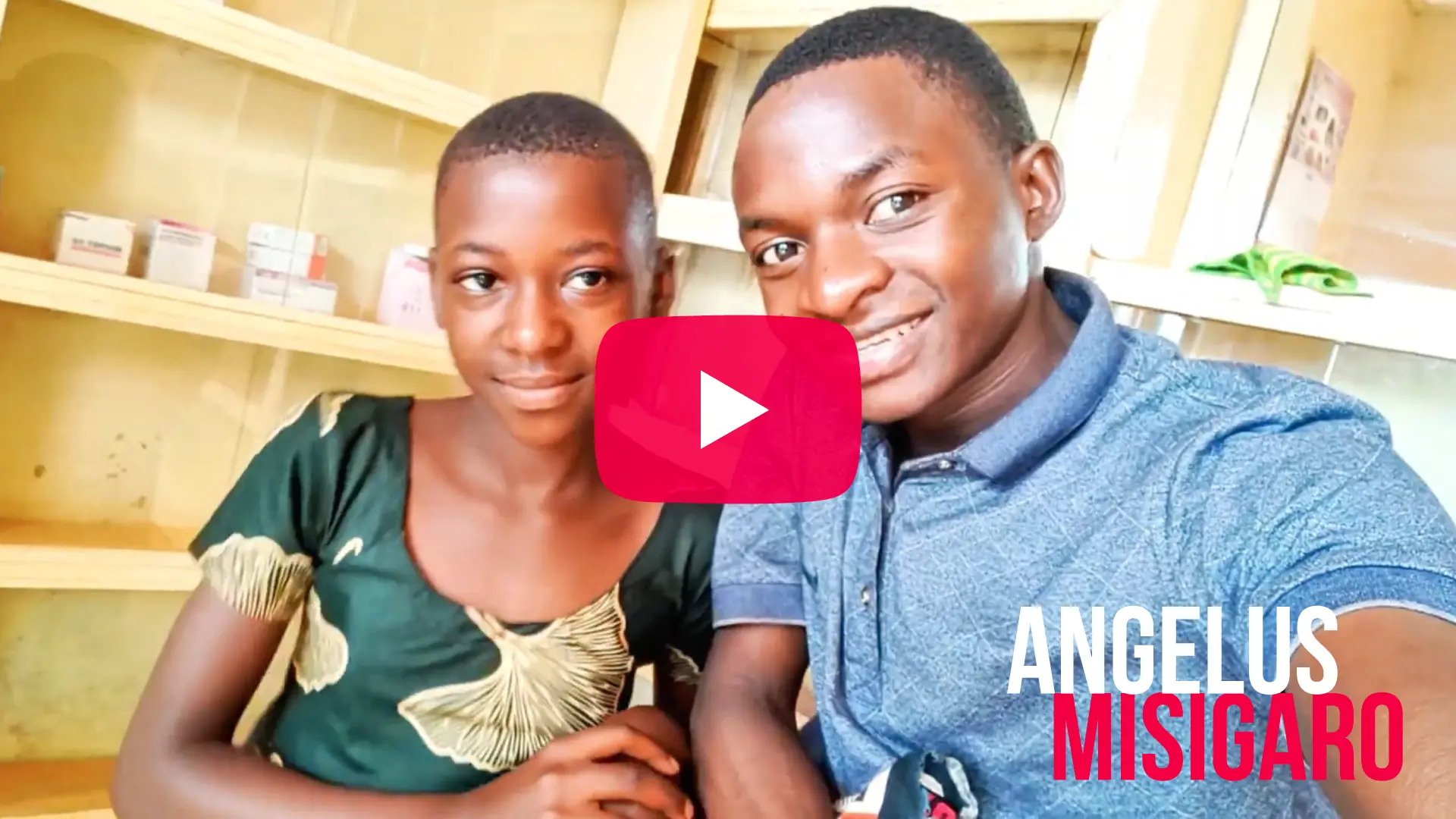 A video thumbnail featuring a smiling young Black man taking a selfie next to a smiling young Black woman. The man is on the right wearing a blue polo shirt, and the woman is on the left wearing a dark green patterned dress. In the center of the image is a red YouTube-style play button overlay. The background shows wooden shelving with a few small boxes on them. In the bottom right corner, the name "ANGELUS MISIGARO" is written in large block letters, with "ANGELUS" in white and "MISIGARO" in bold red.