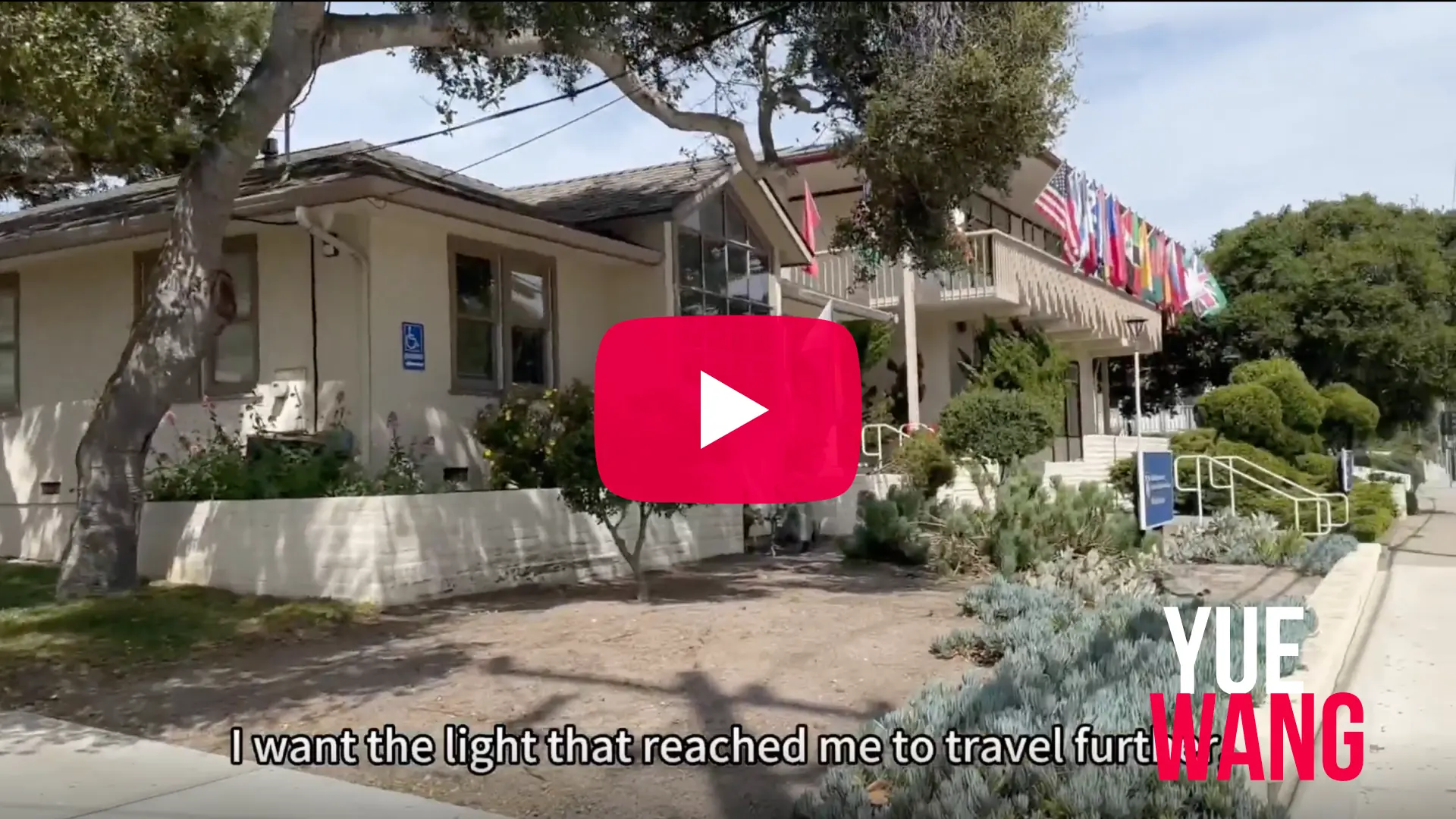 A video thumbnail showing the exterior of a two-story beige building surrounded by green trees and low-growing shrubs under a bright sky. A row of international flags is displayed along the second-floor balcony. In the center of the image is a red YouTube-style play button overlay. At the bottom, a line of white captioned text reads, "I want the light that reached me to travel furt...". In the bottom right corner, the name "YUE WANG" is written in large block letters, with "YUE" in white and "WANG" in bold pink.