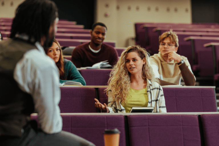 A woman speaks with a professor in a lecture hall surrounded by students, discussing U.S. classroom expectations in an academic setting.