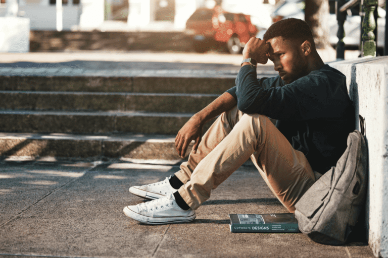 A man sits on a concrete surface outdoors, taking a moment to manage academic pressure and mental health challenges in a campus-like setting.